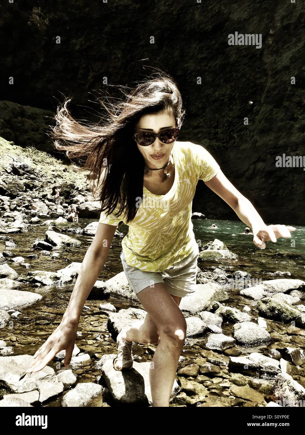 Young woman, long brunette hair, glides gracefully balanced across river rocks - Smartphone Captured Stock Image