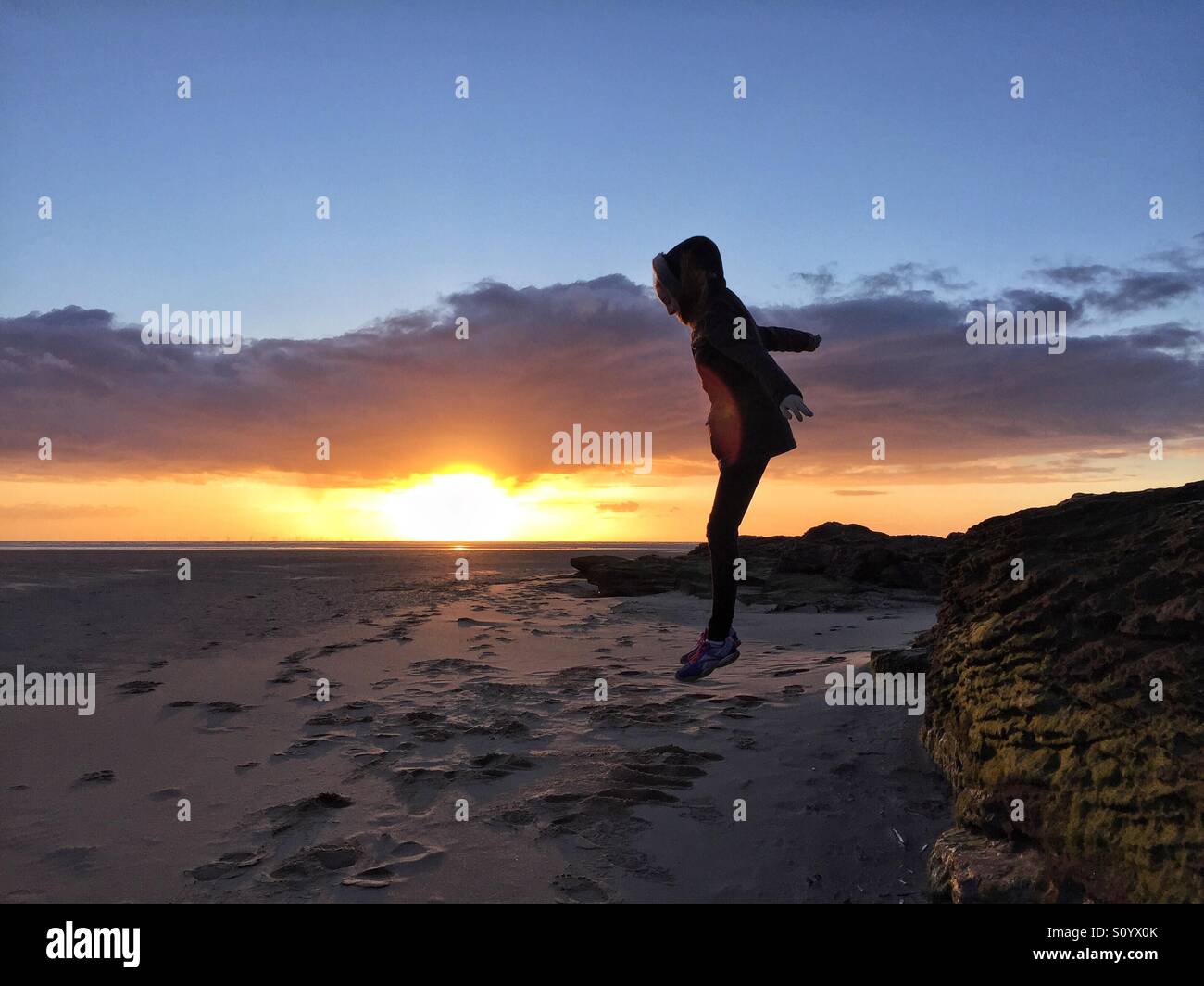 Girl jumping from rocks to sand in front of sunset Stock Photo