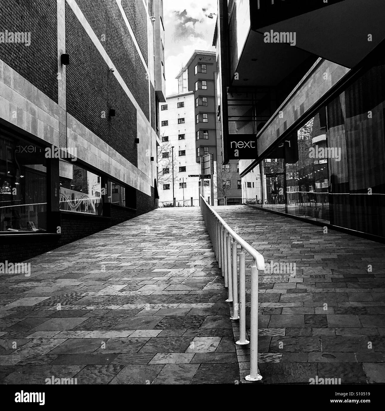 A pedestrian shopping walkway with handrail. Shot in black and white and showing a central vanishing point. The scene is empty, prior to shop opening. Stock Photo