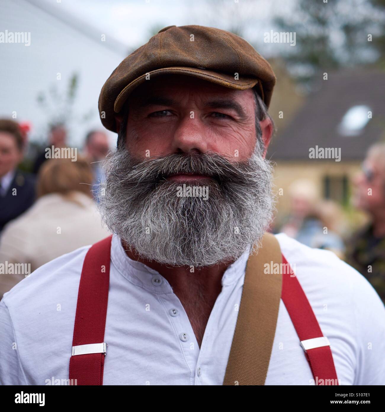 Portrait of a man with a substantial greying beard and flat cap wearing a grandad shirt and red braces. Stock Photo
