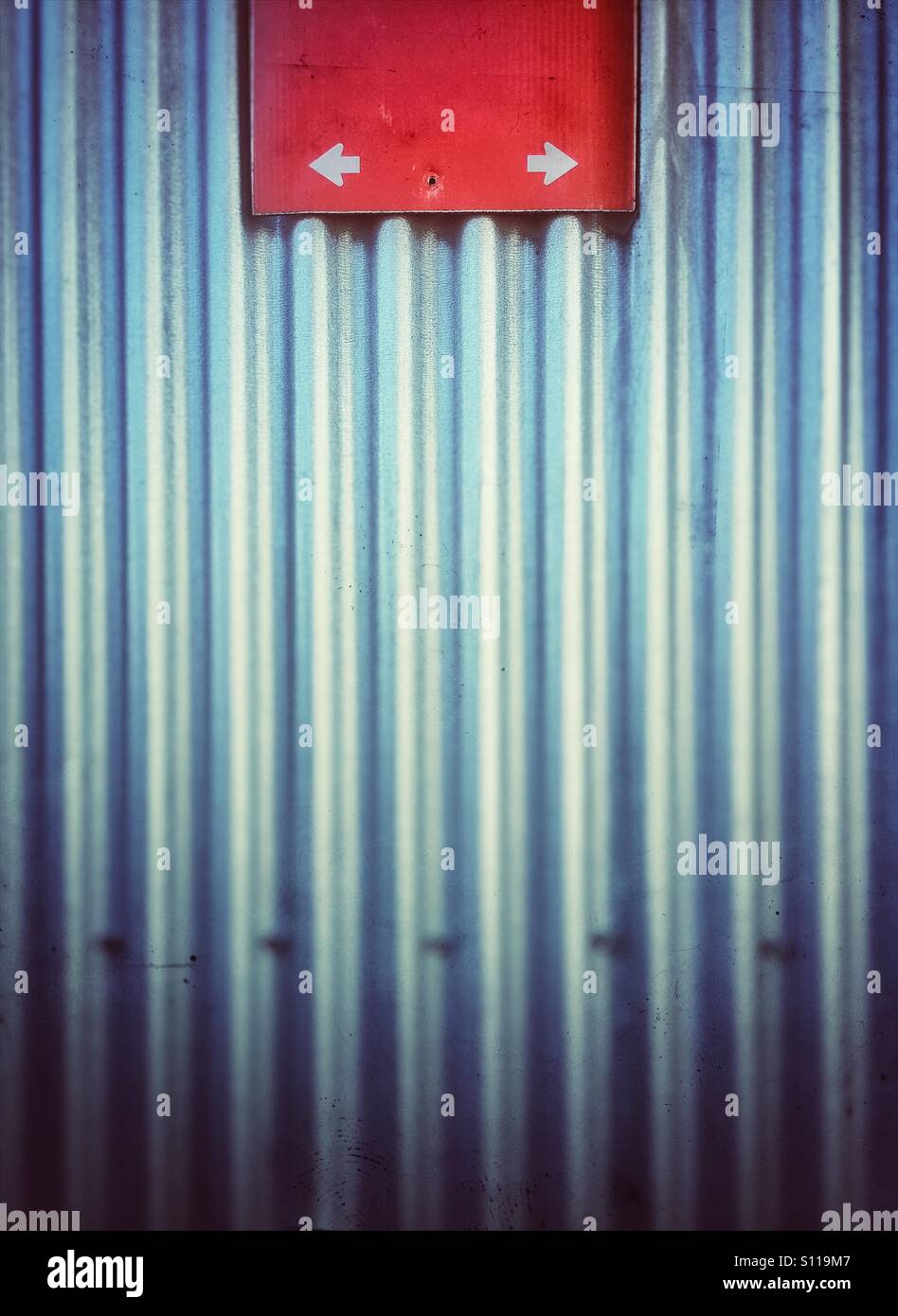 Red sign against a corrugated metal wall with white arrows on sign pointing in opposite directions. - Smartphone Captured Stock Image