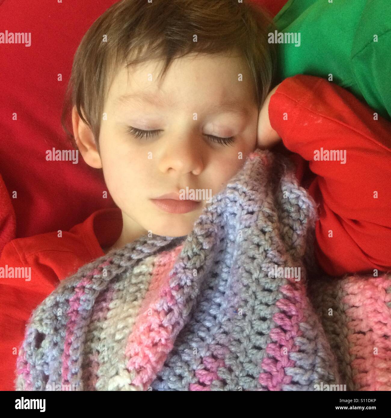 Young boy child sleeping peacefully with a crochet blanket. He is wearing a colourful red t-shirt and is taking his nap on a red and green cushion. He is snuggled up to a handmade crochet throw. Stock Photo