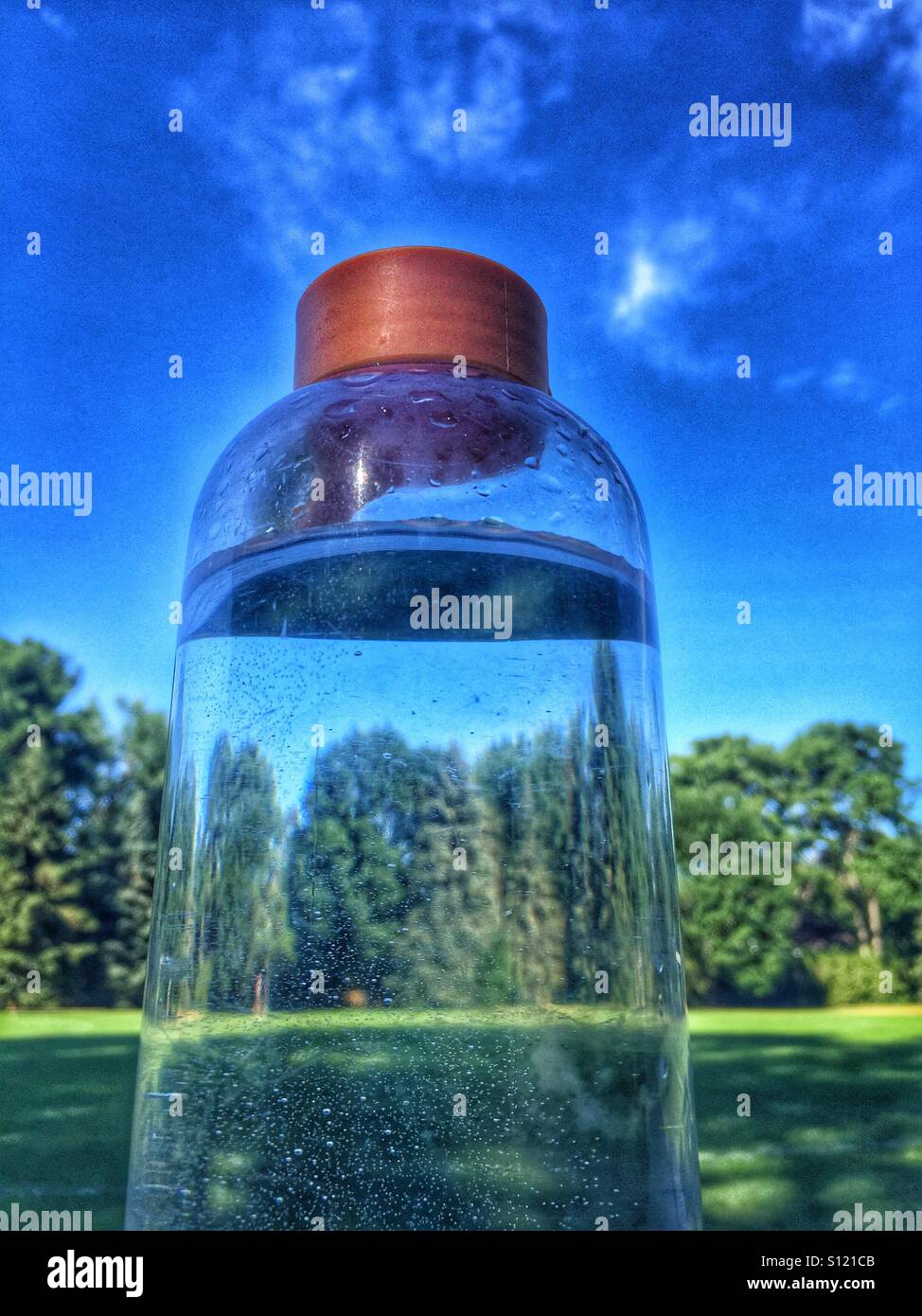 Closeup of clear water bottle in front of grass field, tall trees and blue Sky on a hot summer day in Ontario. - Smartphone Captured Stock Image