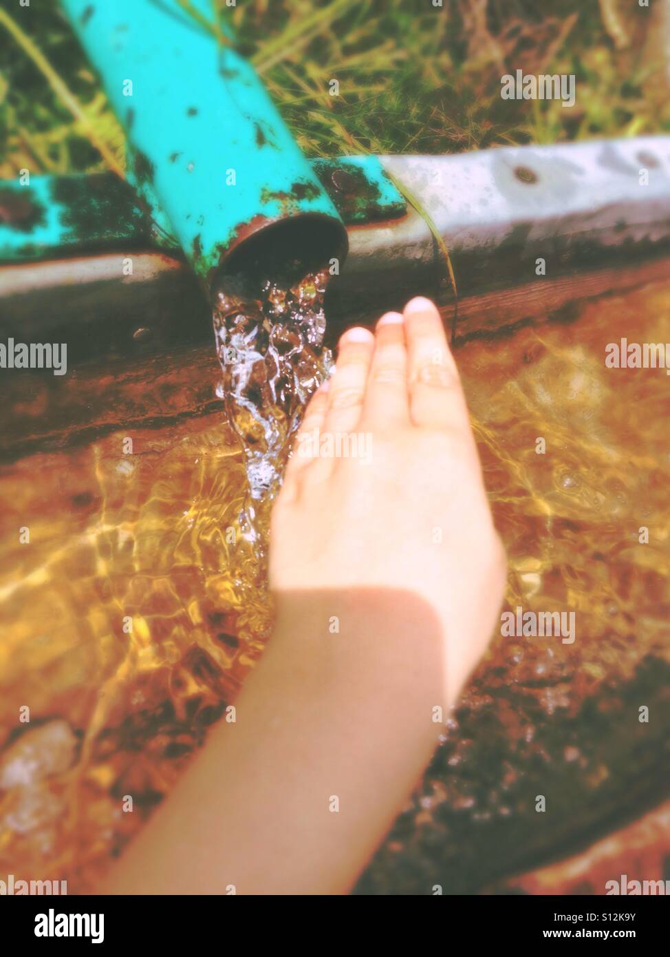 The hand of a caucasian boy reaching for fresh water flowing from a green pipe into a bassin Stock Photo