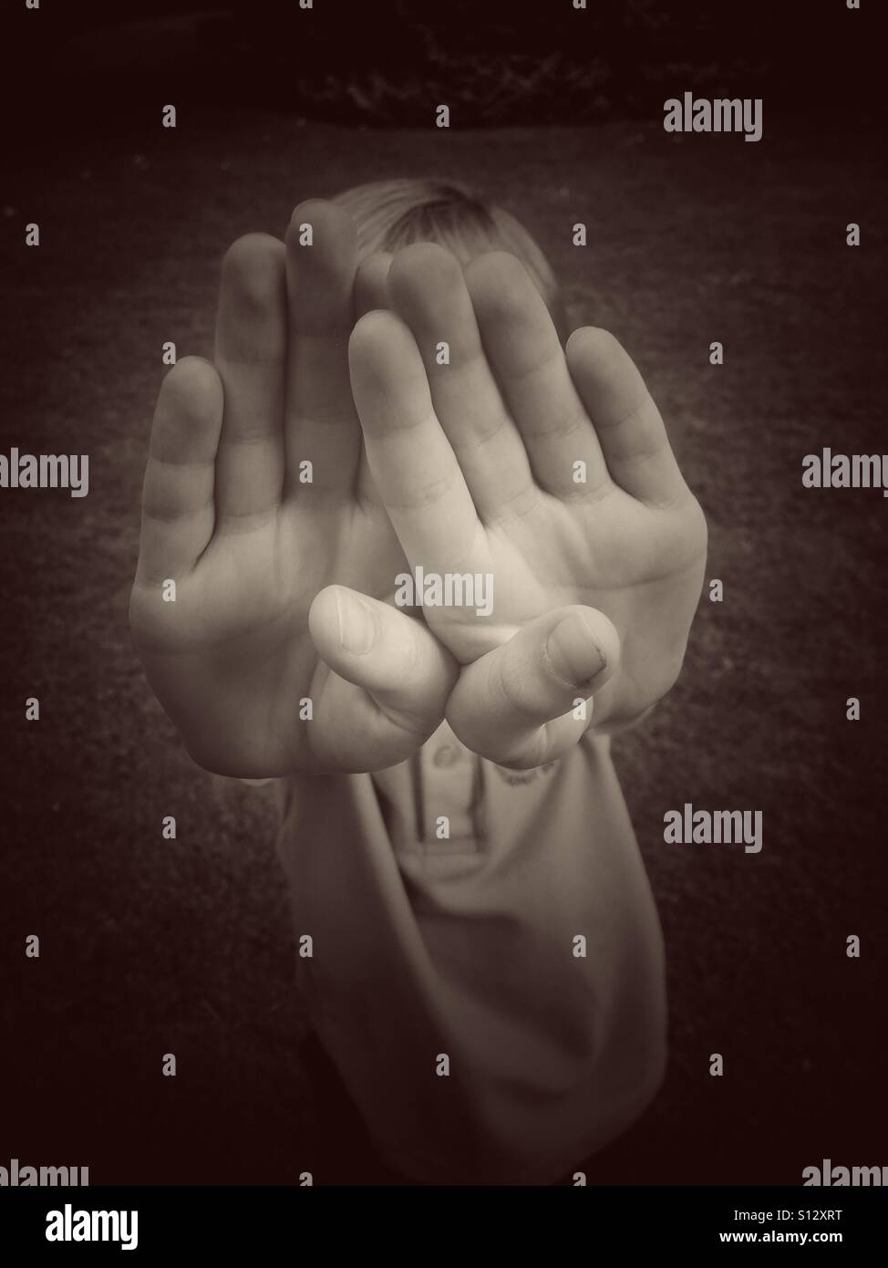 A six year old boy uses both of his hands to cover or protect his face. Is the boy in playful mood OR is he scared of a Paedophile attack? An image to illustrate social issues. - Smartphone Captured Stock Image
