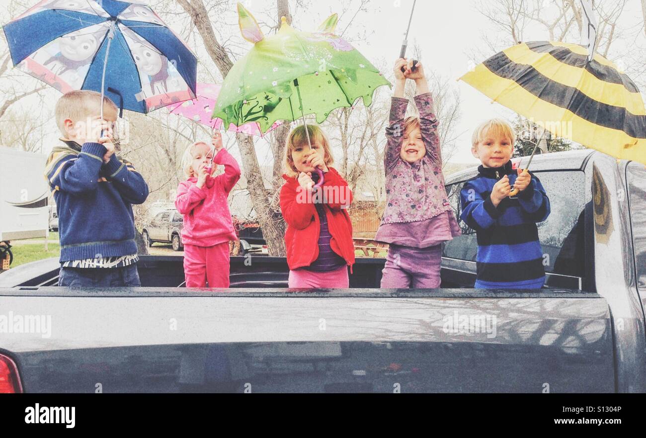 Five children play with umbrellas in the back of a parked pickup truck on a drizzly day. Stock Photo