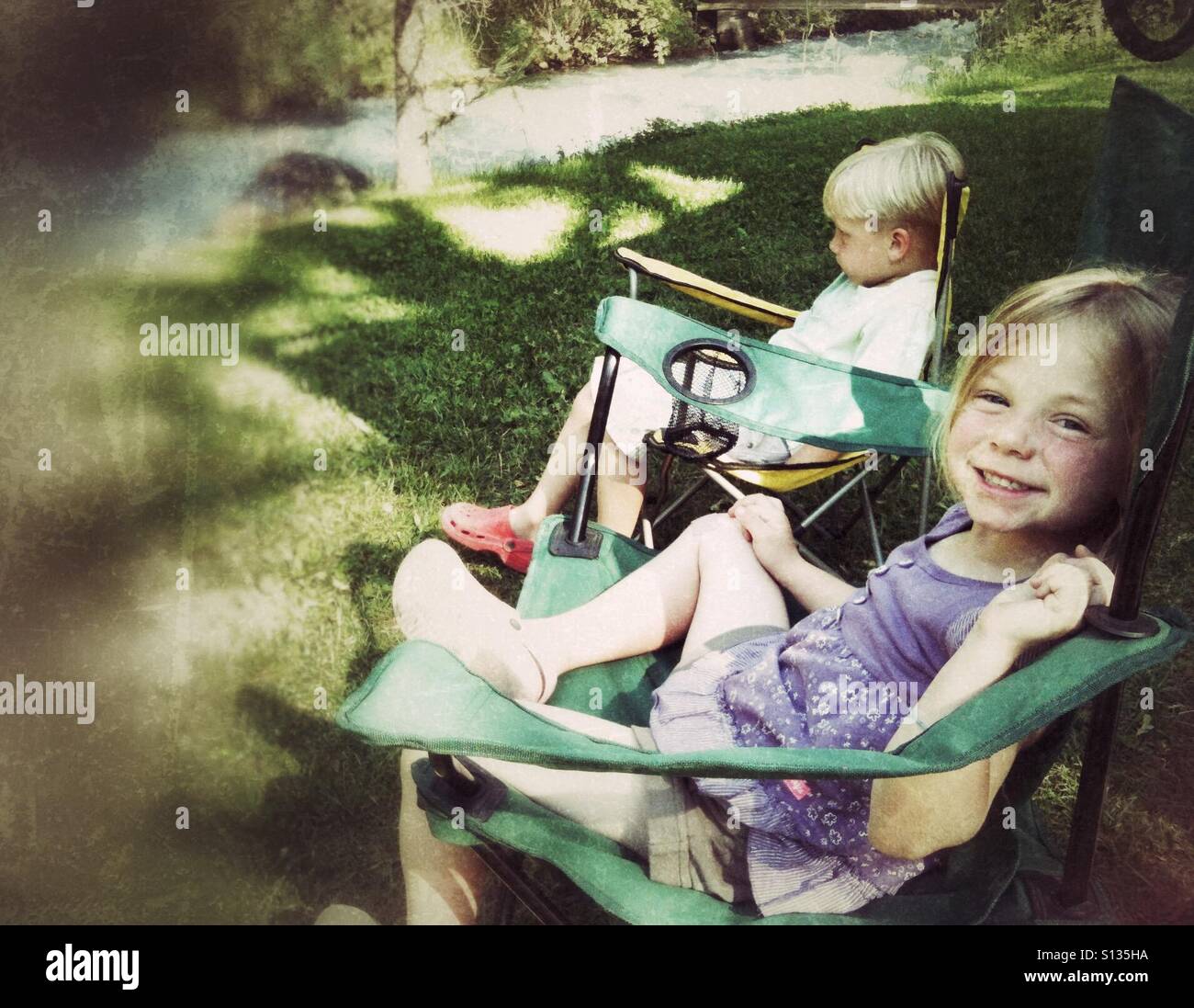 Two children sit in camping chairs beside a creek. - Smartphone Captured Stock Image