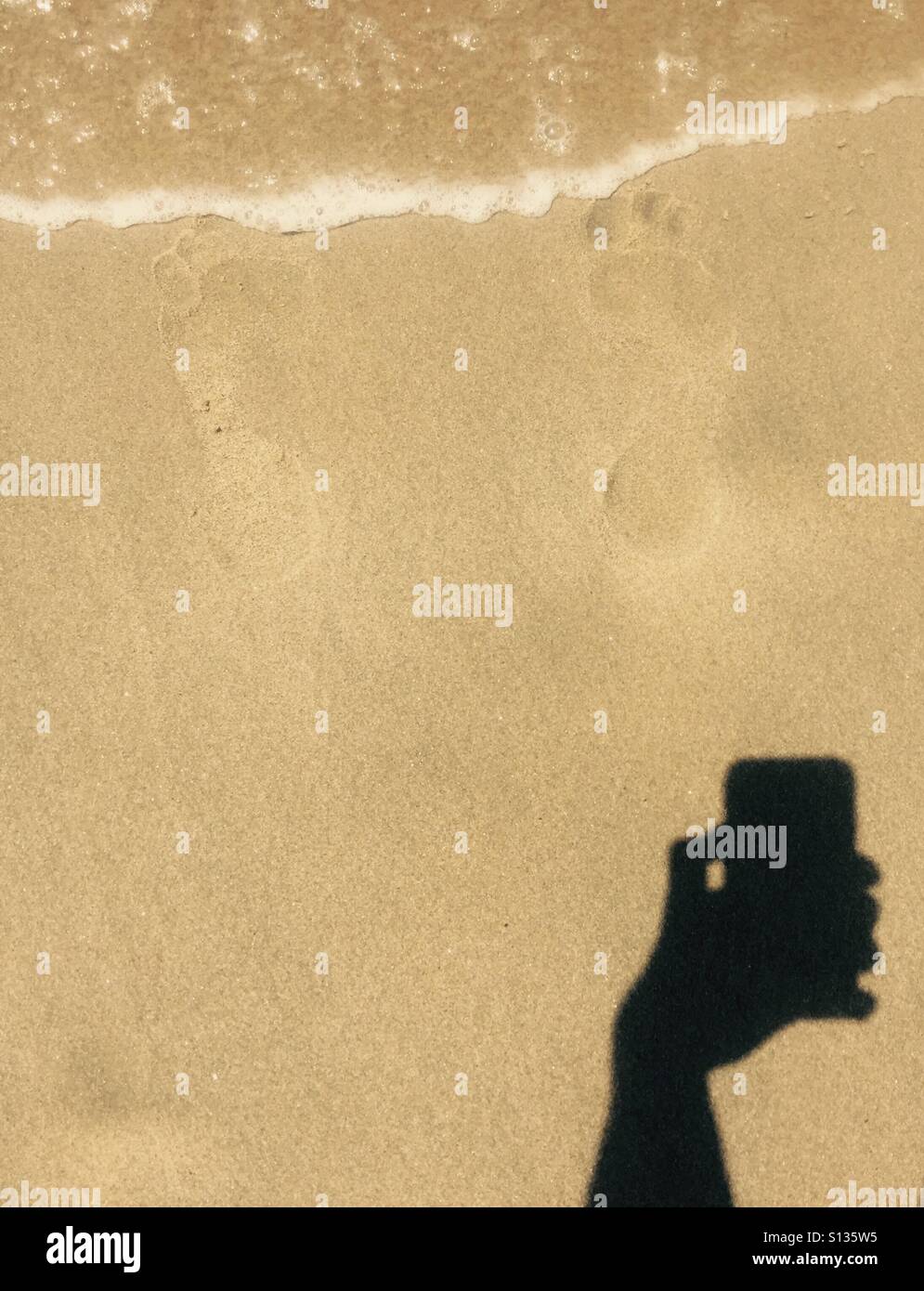A shadow of an hand holding a phone taking a photo of some footprints in the sand. Stock Photo