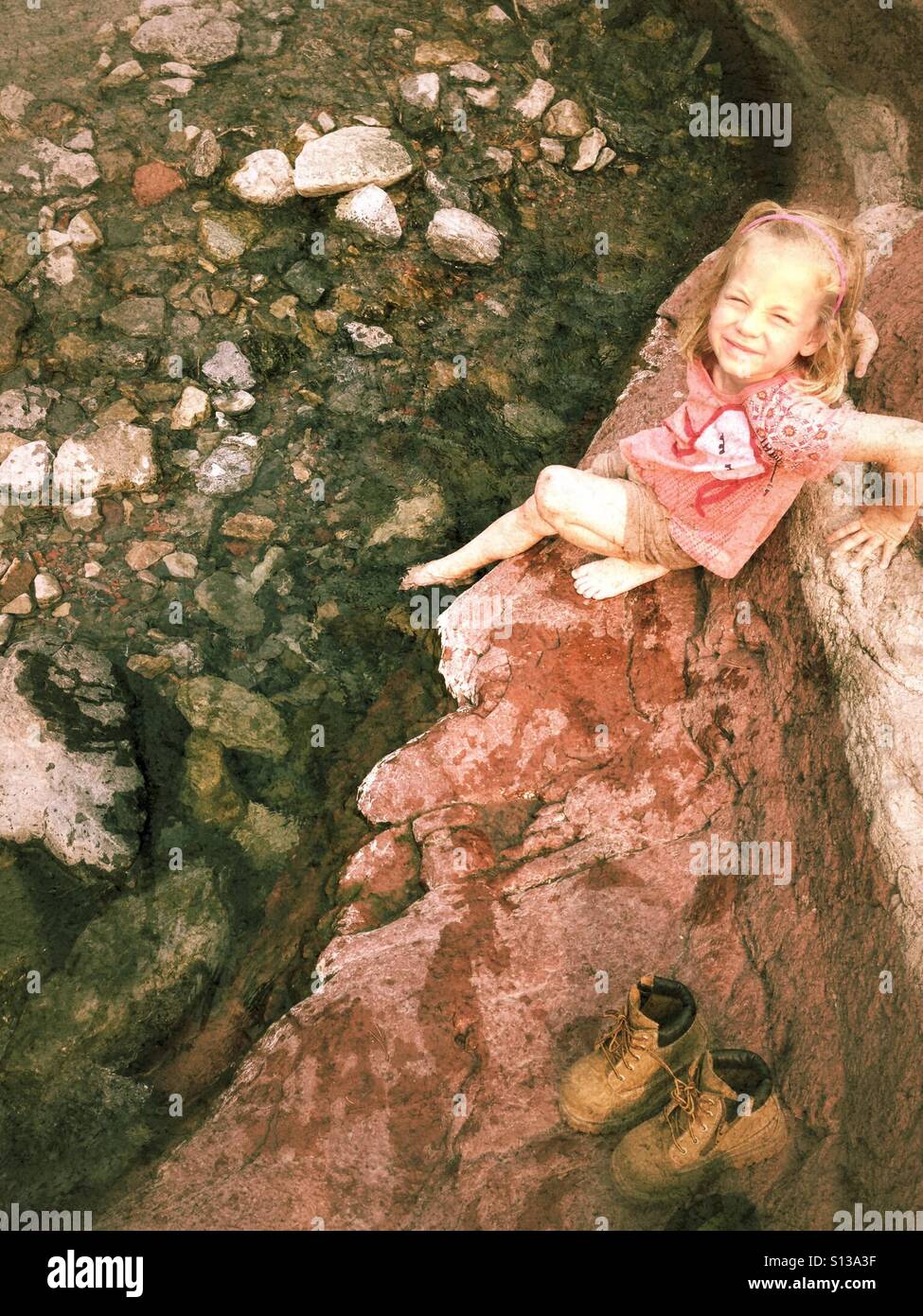 A girl dips her feet in a stream with her hiking boots nearby. - Smartphone Captured Stock Image