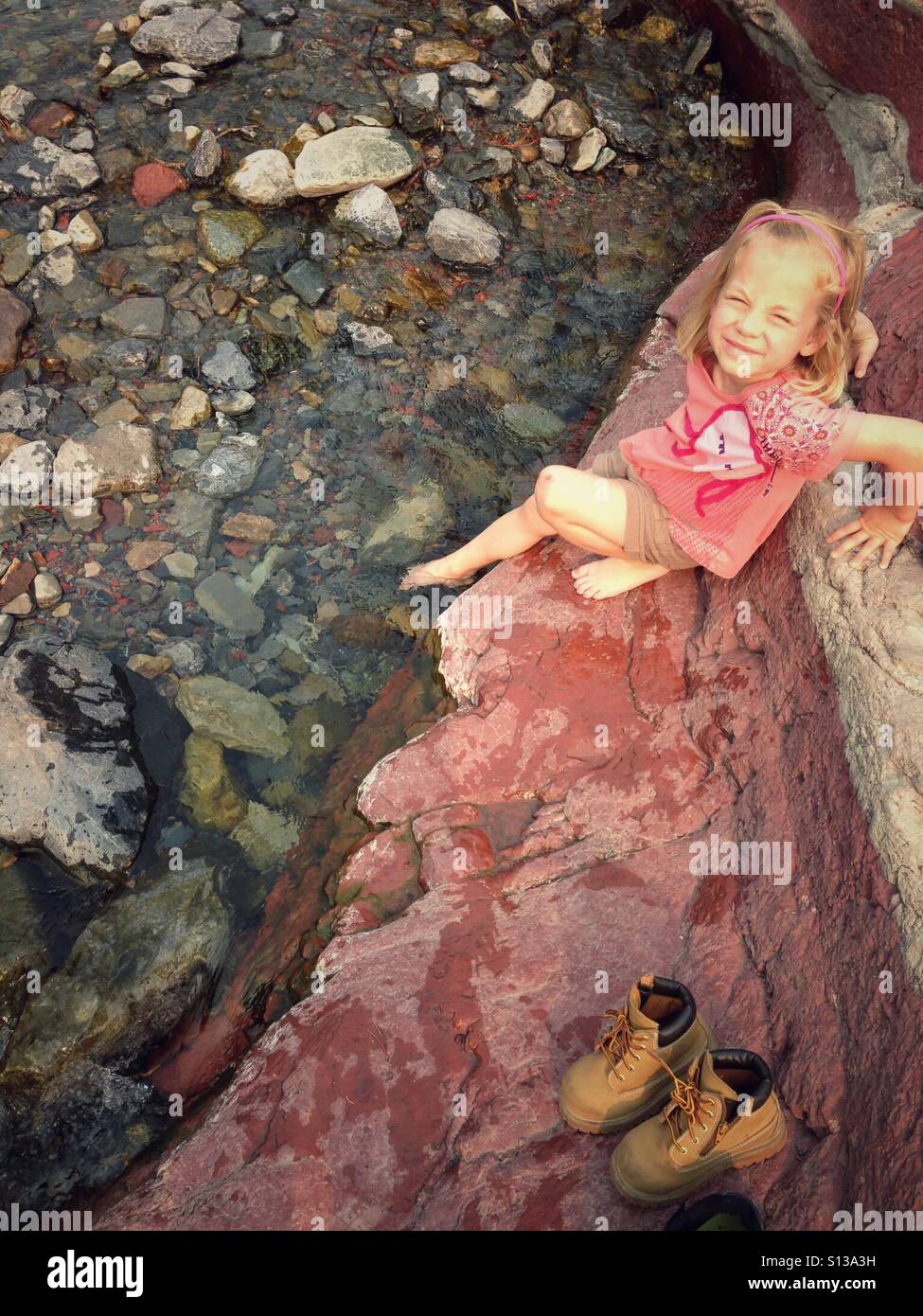 A girl dips a foot into a trickle of water sitting on red rock with her hiking boots nearby. - Smartphone Captured Stock Image