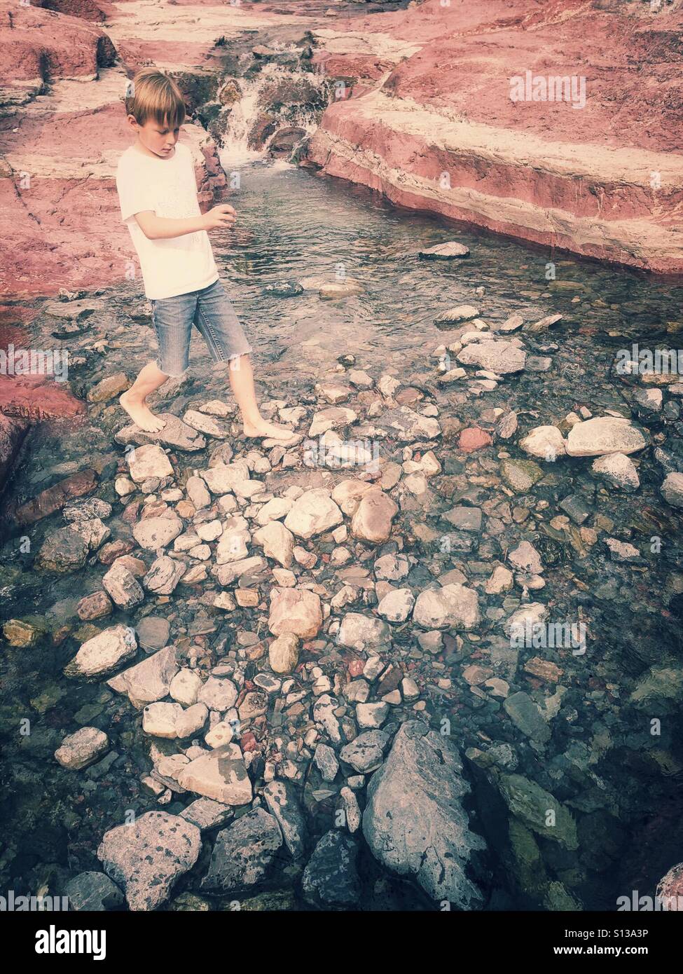 A boy picks his way barefoot across rocks in a stream. - Smartphone Captured Stock Image
