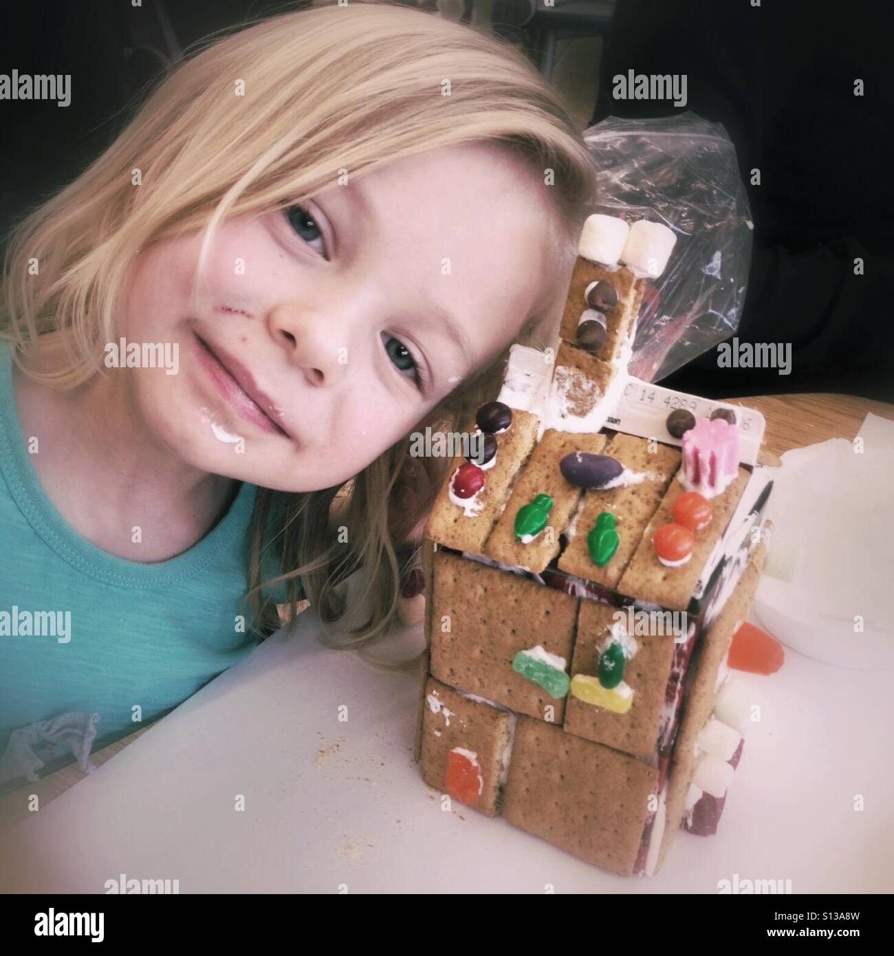 A kindergarten age girl smiles proudly next to a gingerbread house she built. - Smartphone Captured Stock Image