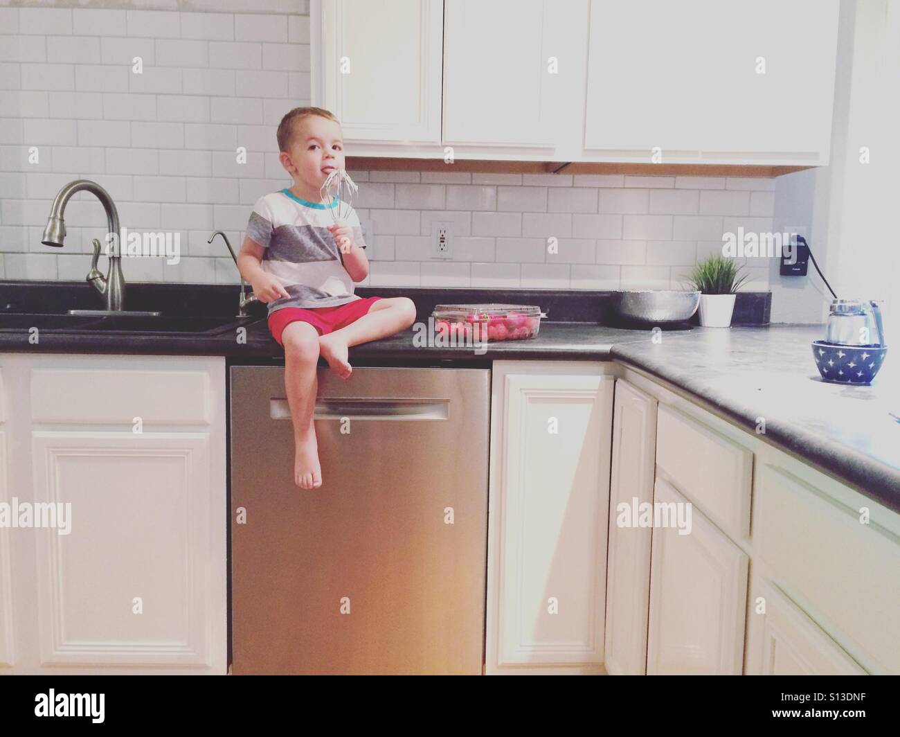 Toddler sitting in the kitchen counter licking whip cream off the beaters Stock Photo