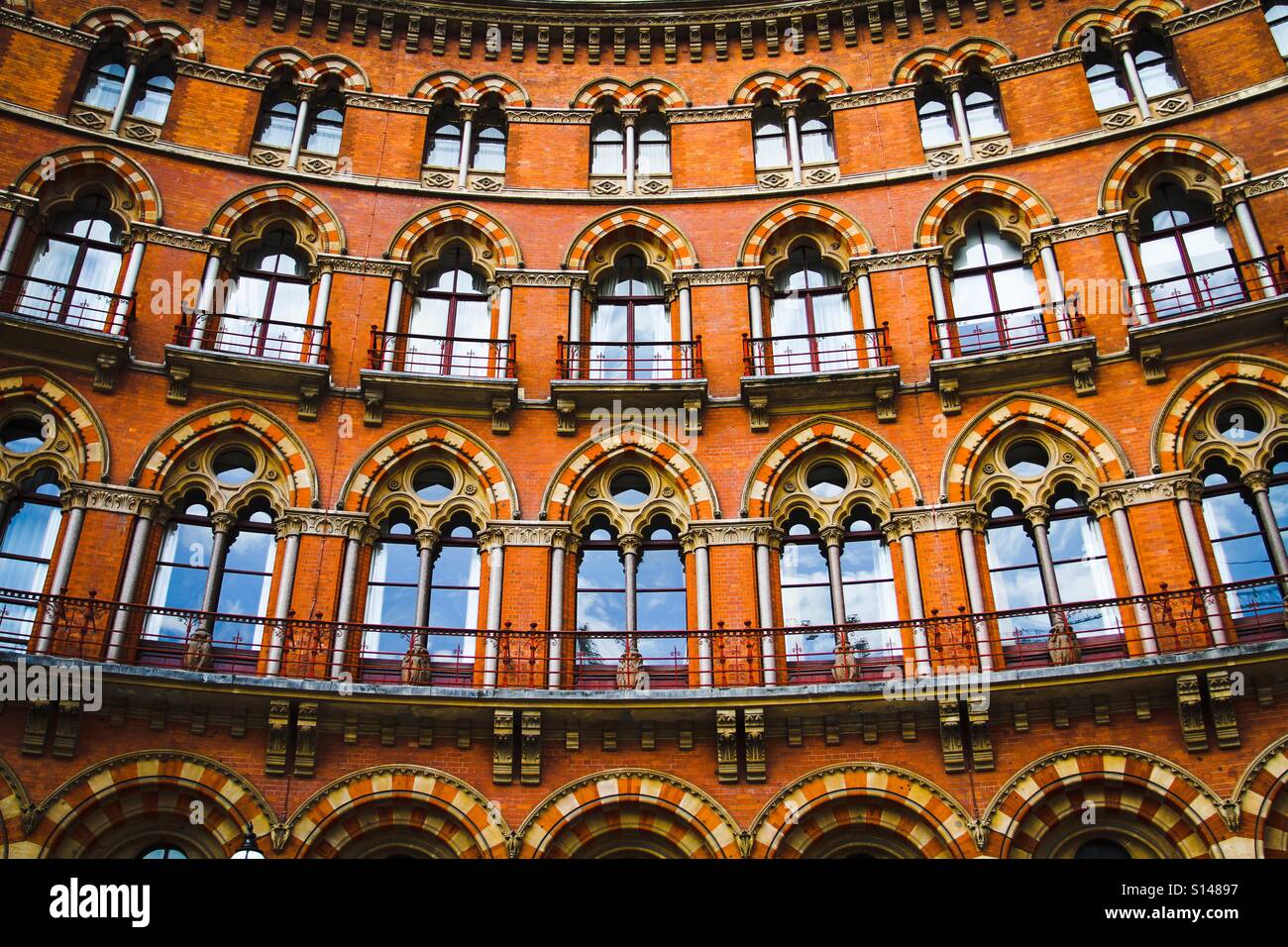 The curved facade of an ornate, Gothic building with rows of uniform, arched windows and elaborate, red brick architecture. Stock Photo
