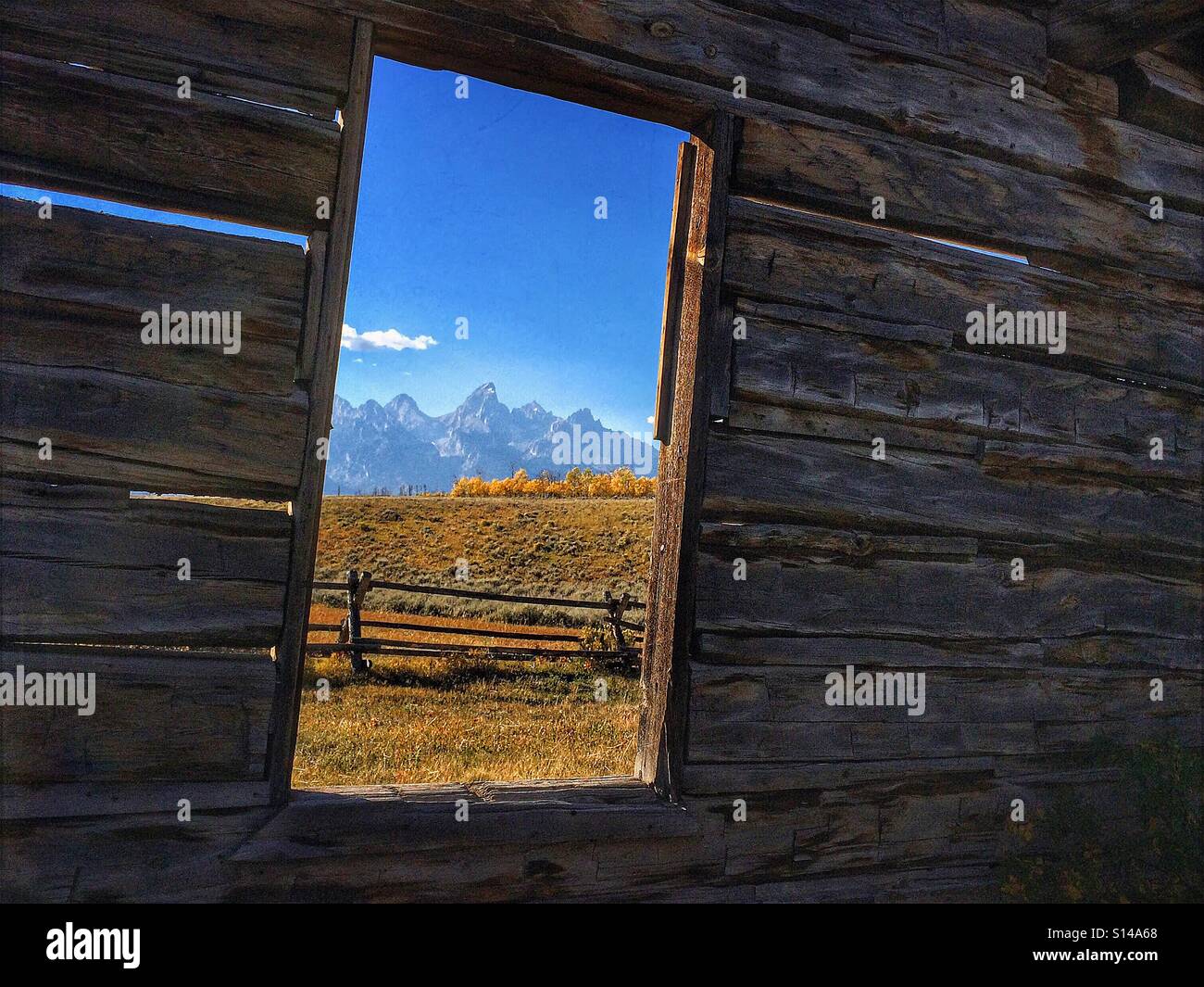 View of the Tetons and Grand Teton National Park and rail fence through the window of an old abandoned log cabin. - Smartphone Captured Stock Image