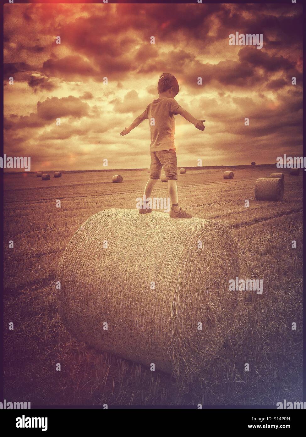 A retro film effect image of a 7 year old boy balancing on top of a Hay Bale. The location is a recently harvested field in the South of England. Photo Credit - © COLIN HOSKINS. - Smartphone Captured Stock Image