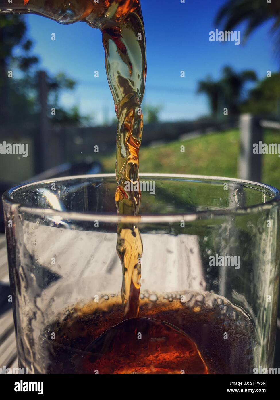 Soft drink being poured into a glass with ice with tropical cal for plants in the background Stock Photo