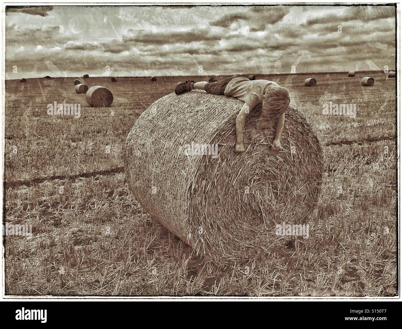 A seven year old boy lies on a freshly produced hay bale in a field that has recently been harvested. Is the boy asleep? Is he playing? Where are his friends? Parents? Photo Credit - © COLIN HOSKINS. - Smartphone Captured Stock Image