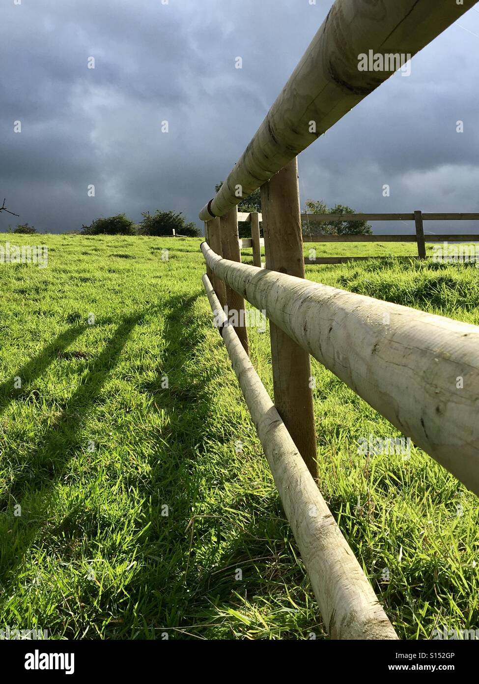 Post and rail fence in a field with shadows from the afternoon sun and storm clouds behind Stock Photo