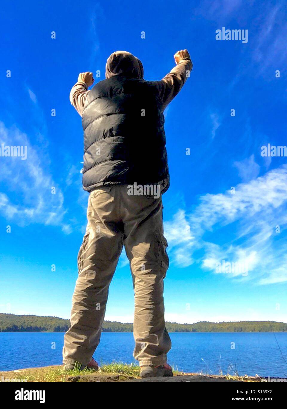 Person At Sunny Lakeshore With Arms Up Pointed Towards The Sky Stock Photo