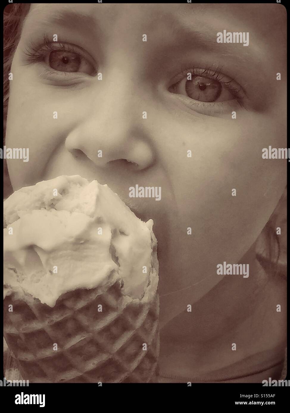 A close up, sepia toned image of a 4 year old girl eating a large cone filled with Vanilla Ice Cream. The joys of childhood and summer! Photo Credit © COLIN HOSKINS. - Smartphone Captured Stock Image