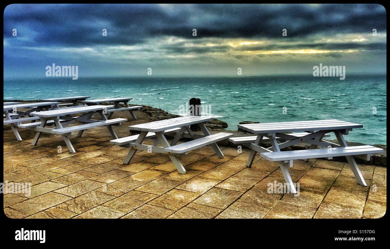 A selection of abandoned wooden seats and tables overlooking the Atlantic Ocean. The sun is starting to set on a cold and windy winters day. Photo Credit - © COLIN HOSKINS. - Smartphone Captured Stock Image