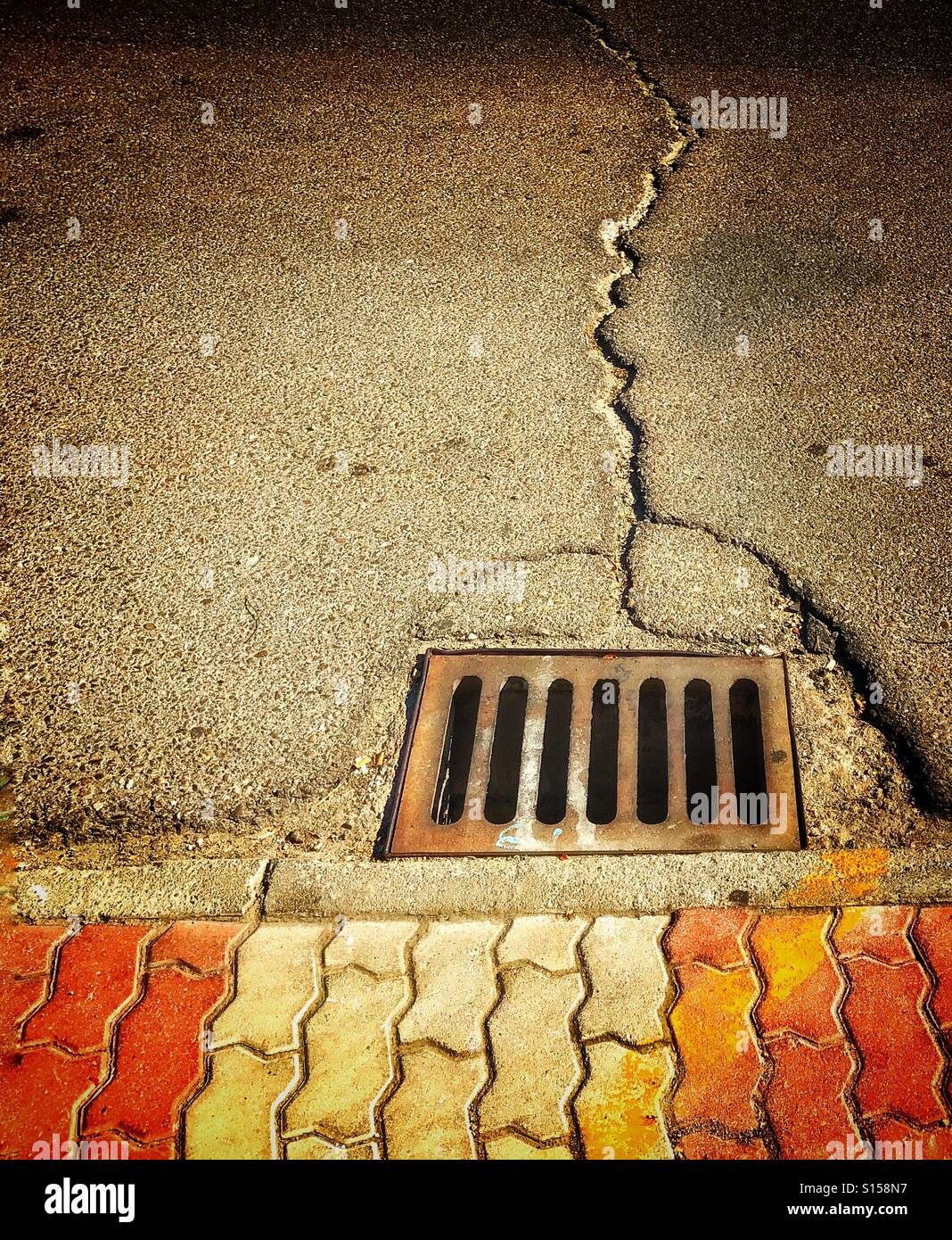 Drain next to ornate tiled pavement surrounded by cracked tarmac of road surface Stock Photo