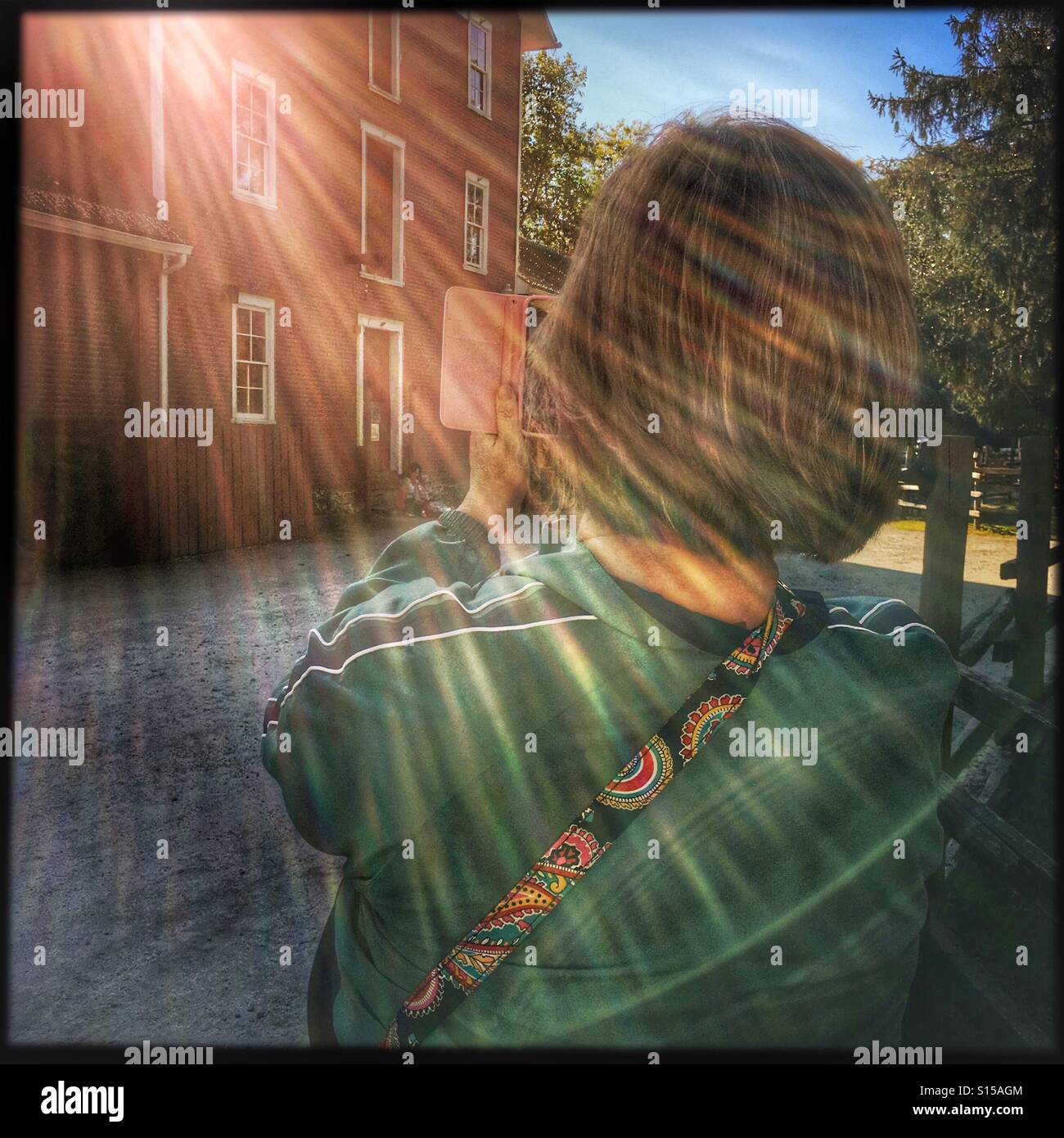 A lady taking a photo of a red building with an Iphone.  The sun creates prism-like rays across the scene. - Smartphone Captured Stock Image A lady taking a photo of a red building with an Iphone.  The sun creates prism-like rays across the scene. - Smartphone Captured Stock Image
