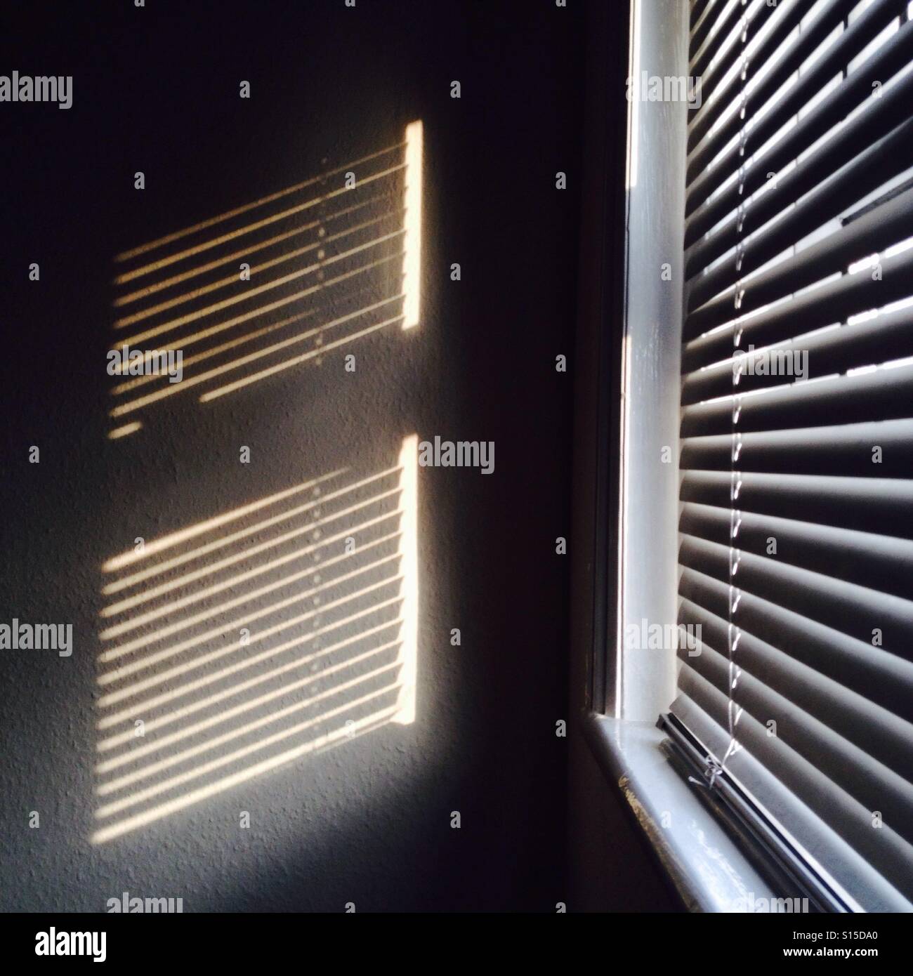 Sunlight seeping through a closed window blind casting a reflection on woodchip wallpaper. Stock Photo