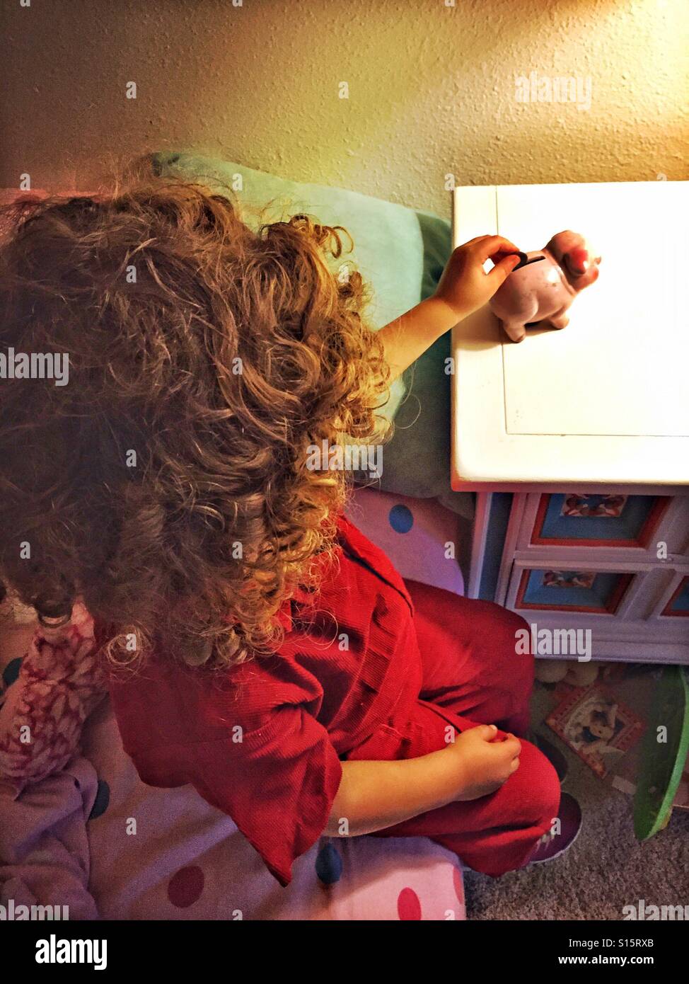 A little girl sits on her bed and deposits a coin into the piggy bank on her bedside table. Stock Photo