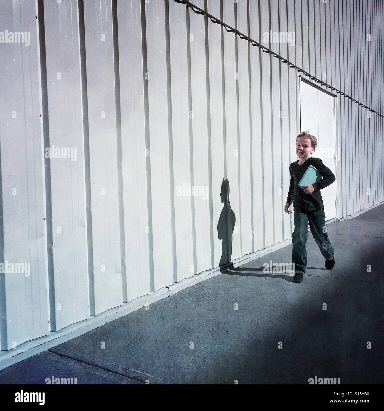 A boy with his tablet under his arm skips beside a commercial building. - Smartphone Captured Stock Image