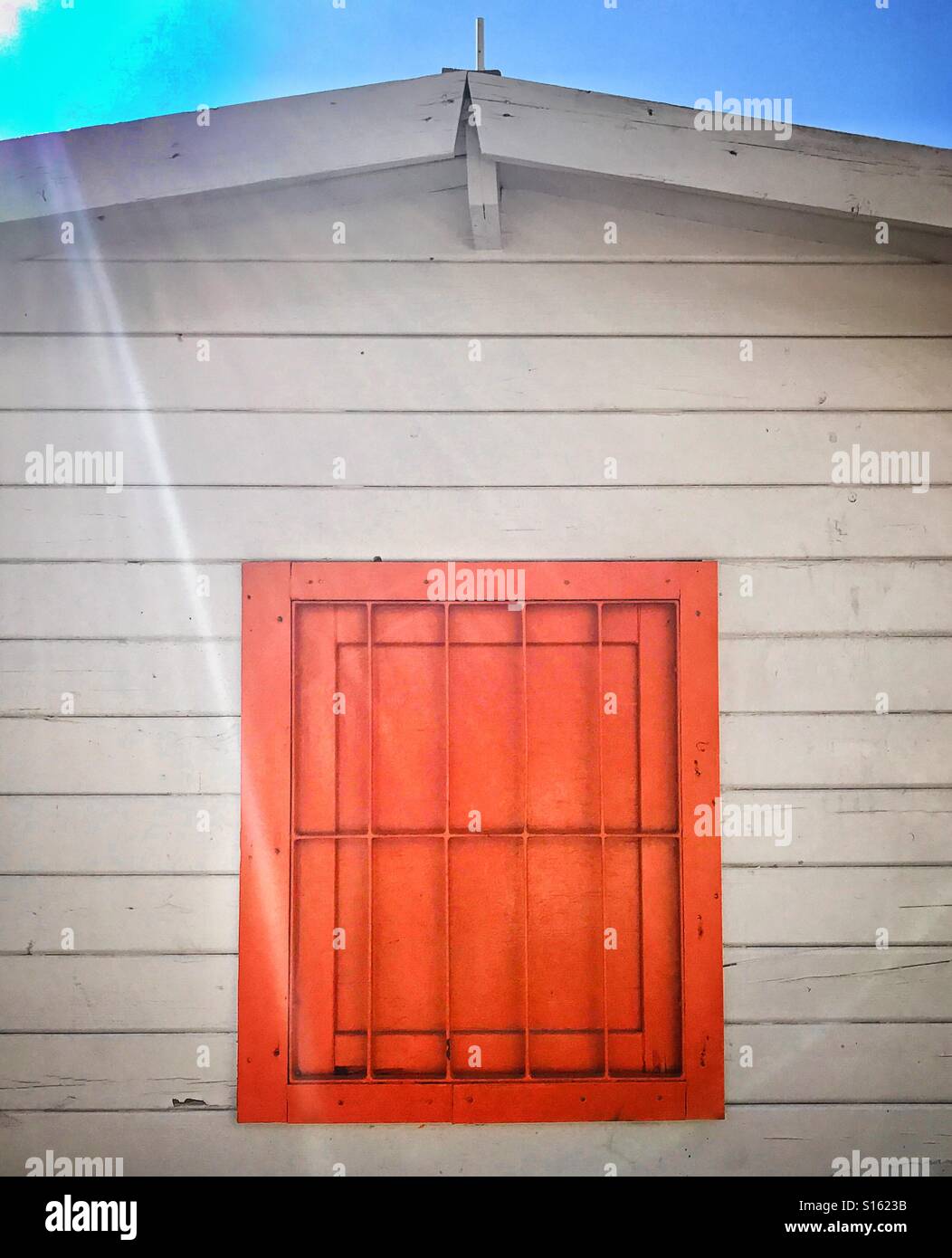 Exterior of wooden beach hut painted white with red window protected by metal security bars under blue sky - Smartphone Captured Stock Image