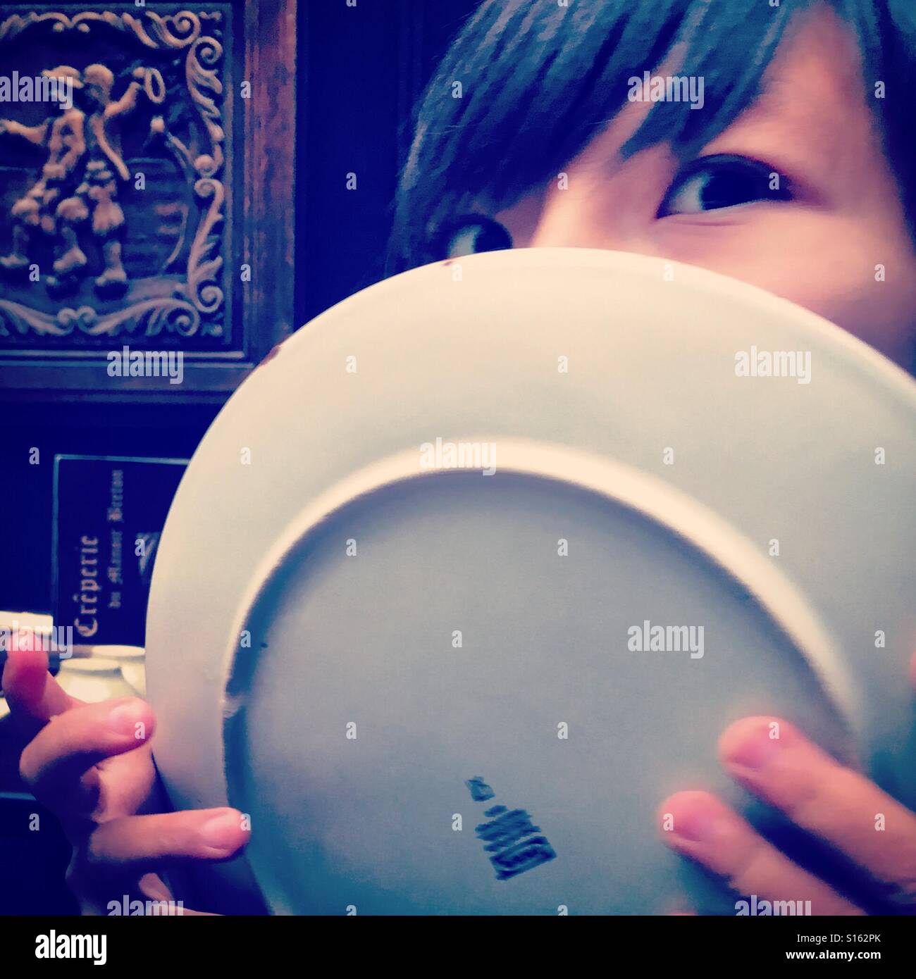 Kid licking its plate afyer finishing his meal lunch diner as it was so yummy tasty nice food to eat eating hungry greedy big eyes half asian kid boy - Smartphone Captured Stock Image