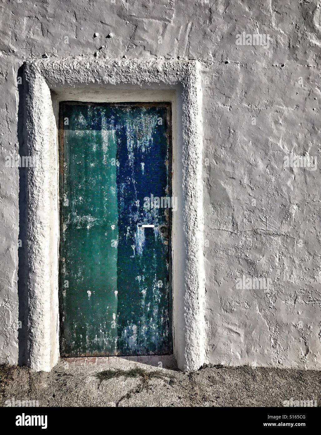 Heavily weathered door painted blue and green set with one rough cast brilliant white concrete wall of sea facing property on the coast of southern Spain near Caleta de Velez. - Smartphone Captured Stock Image