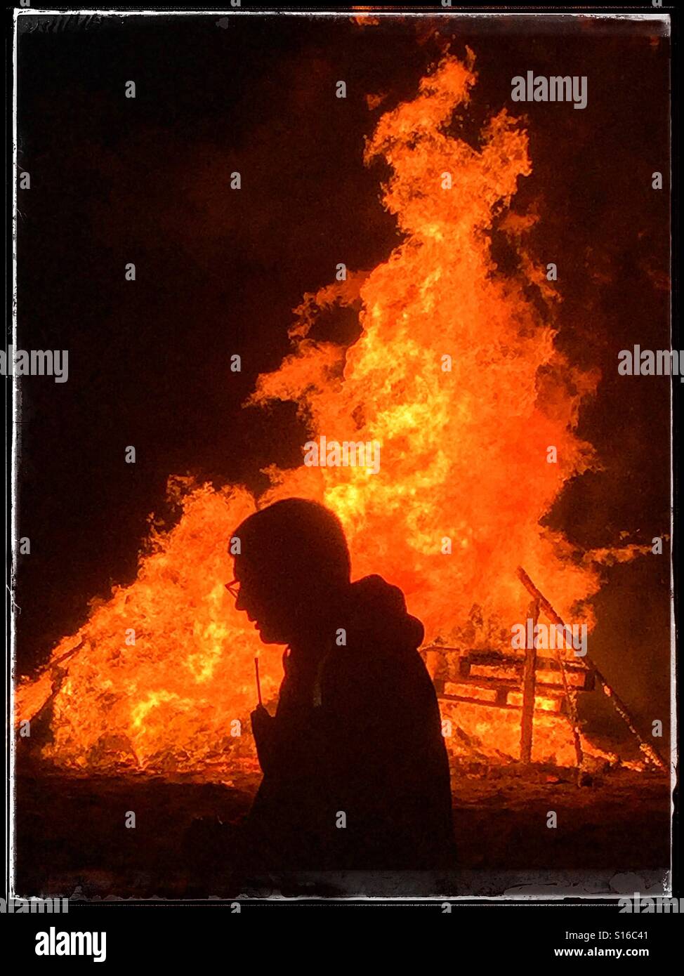 The silhouette of a man as he walks past a raging bonfire during 5th November Celebrations (Guy Fawkes & Bonfire Night). Wooden pallets are burning in an intense fire. Photo Credit © COLIN HOSKINS. - Smartphone Captured Stock Image