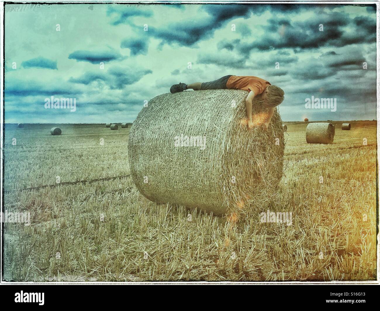A 7 year old boy lies on top of a newly harvested Hay Bale. His arms are hanging over the Bale's edge. Is he asleep? Is he playing a game? Is he alone? Should he be at school? Photo © COLIN HOSKINS. - Smartphone Captured Stock Image