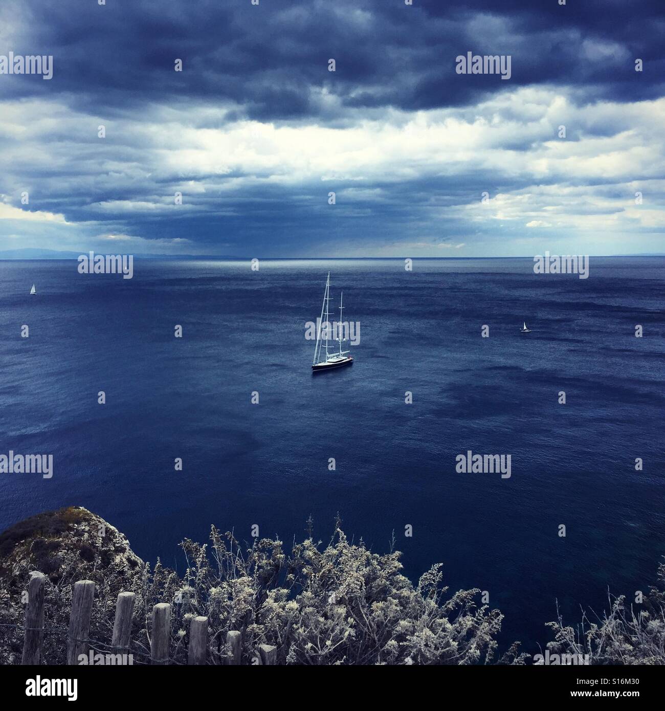 Looking out over the calm ocean at a yacht floating on the horizon under dramatic clouds Stock Photo
