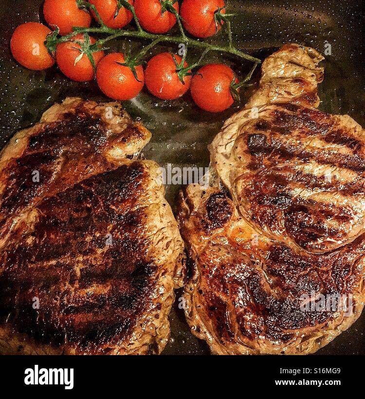 2 Rib eye steaks cooking on a griddle pan with vine tomatoes Stock
