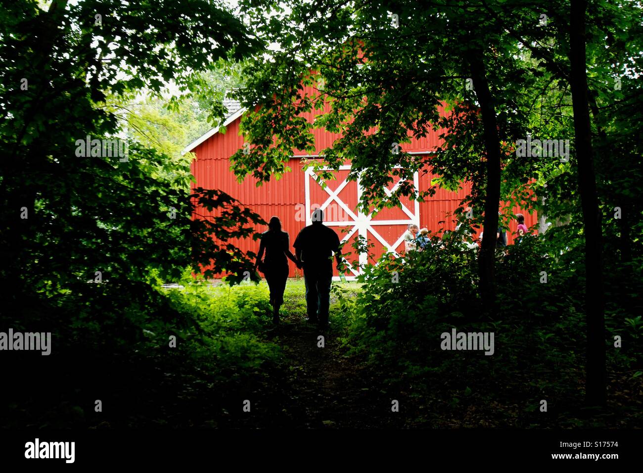 A couple walks out of a wooded area into a clearing where there is a red barn - Smartphone Captured Stock Image