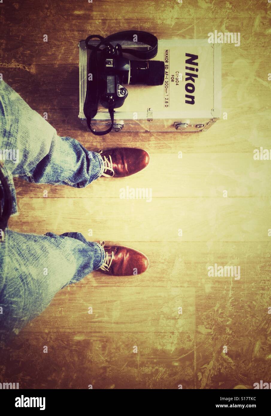 Man wearing brown leather shoes is standing on a oak floor next to him is a Nikon retro flightcase with on top of it a modern Nikon camera with lens - Smartphone Captured Stock Image