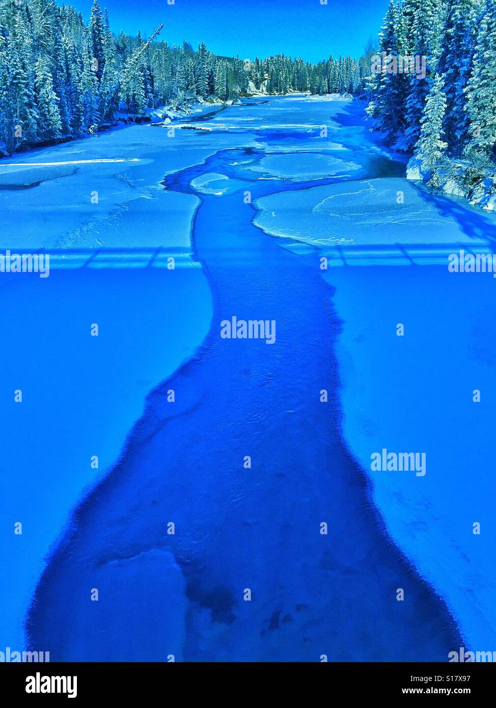 Frozen River viewed from a bridge with bridge shadow and lined by evergreens - Smartphone Captured Stock Image