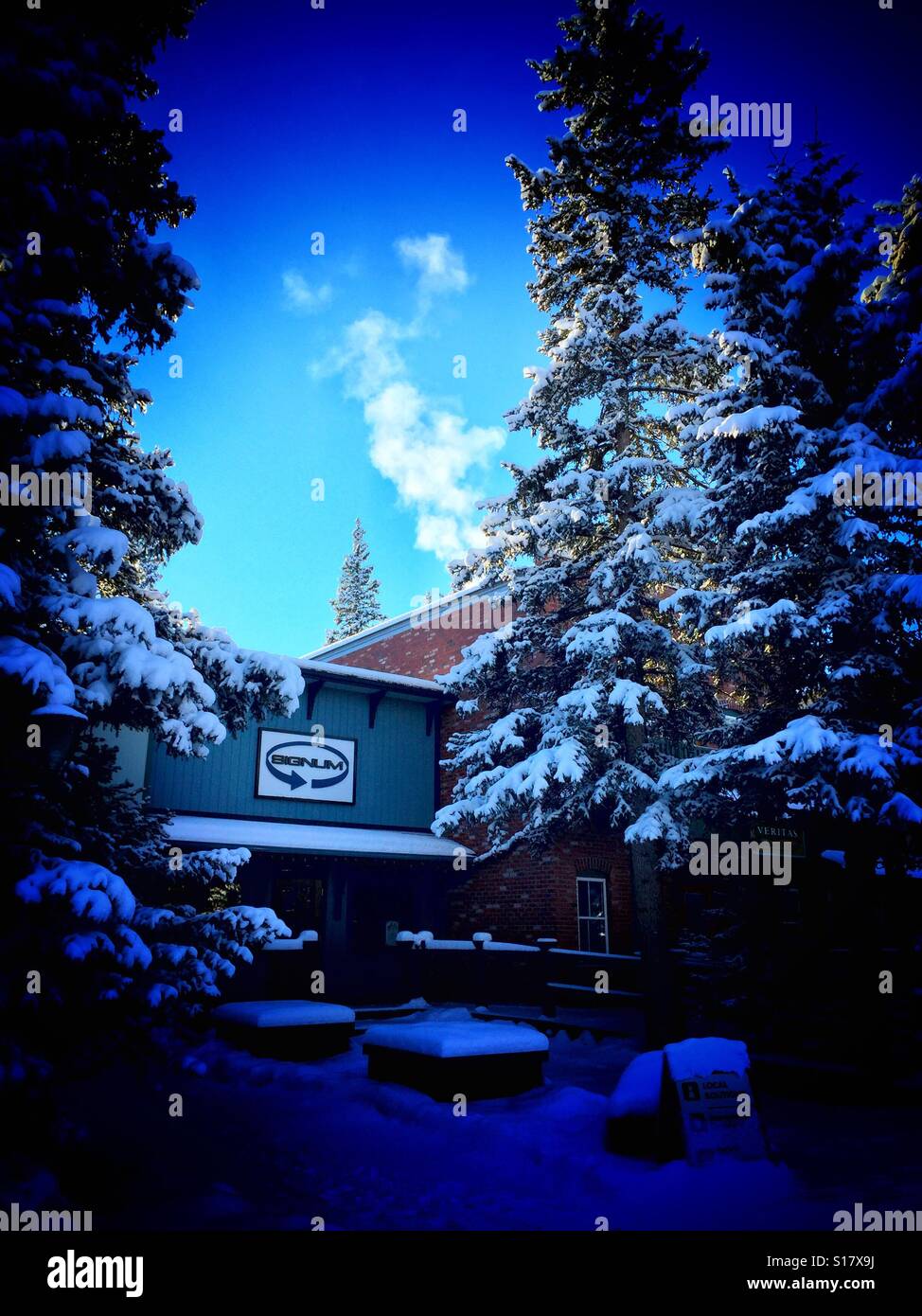 Bragg creek strip mall surrounded by snow covered evergreens with exhaust from one of the businesses - Smartphone Captured Stock Image