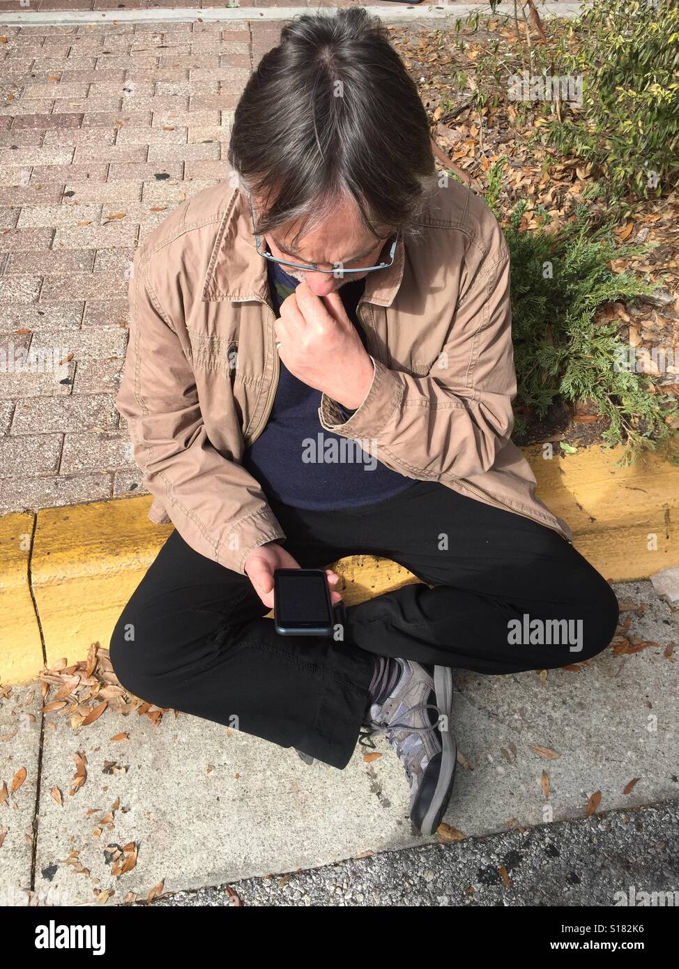 Man sitting on sidewalk curb looking at his phone Stock Photo