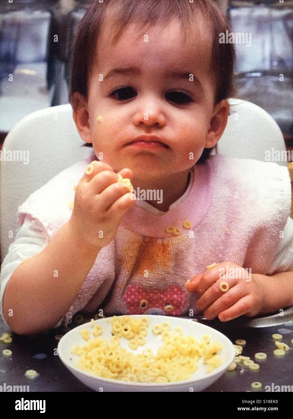 An infant girl in a high chair messily eating noodles Stock Photo
