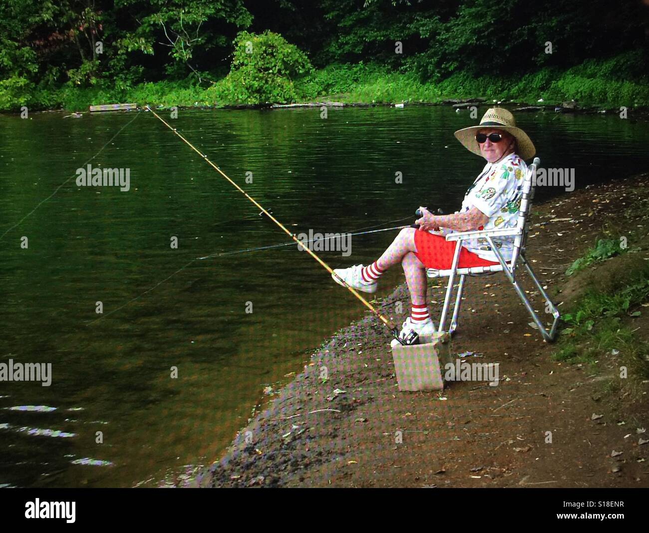 An elderly woman dressed in shorts and a hat sitting in a lawn chair fishing at a lake Stock Photo