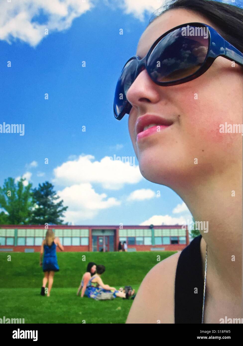 A close up profile of a dark haired girl wearing dark sunglasses against a blue sky with clouds and a school and schoolyard in the background - Smartphone Captured Stock Image