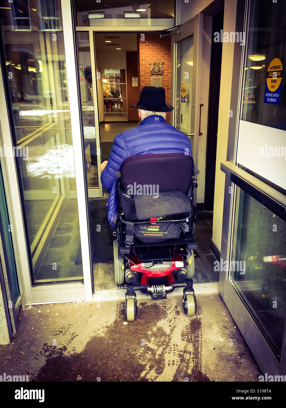 Wheelchair man, mobility challenged, entering a modern building, Canada. Senior. Grey hair. Hat. - Smartphone Captured Stock Image