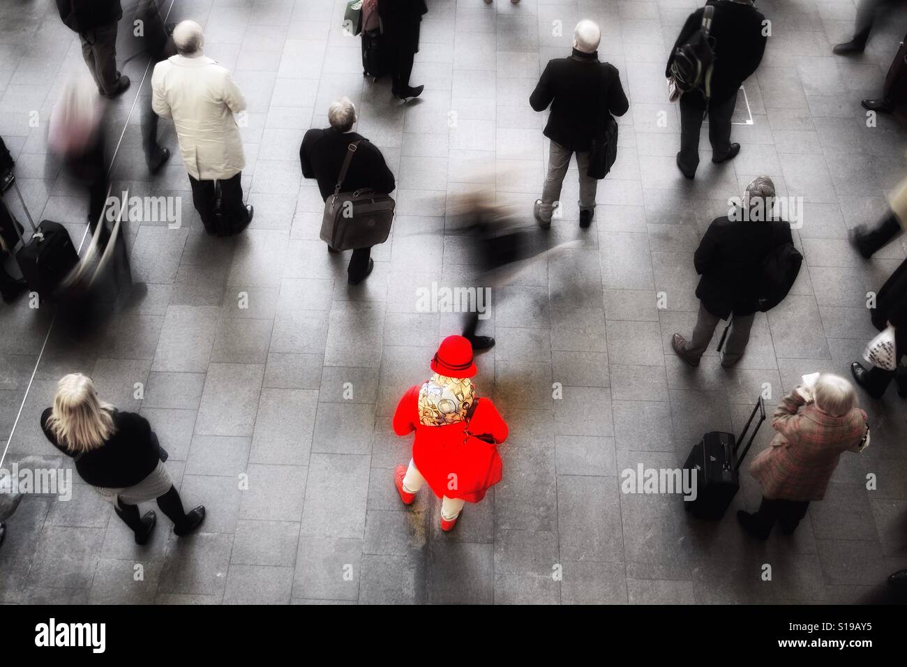 Looking down from above onto a busy train station concourse with a lone woman in red standing out from a crowd of rail commuters and passengers. - Smartphone Captured Stock Image