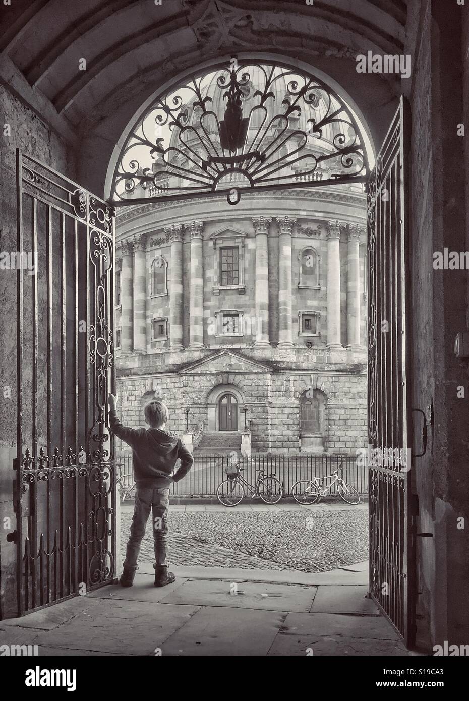 A 7 year old boy stands in an archway of Divinity College & looks at the Radcliffe Camera Building in the University area of Oxford, England, UK. What could the boy be thinking about? © COLIN HOSKINS. - Smartphone Captured Stock Image