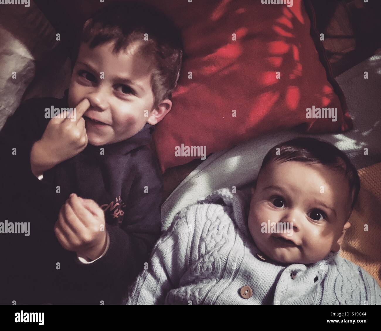 Two brothers lying down next to each other on pillows, one of the boys is picking his nose and the other is just looking straight to camera. - Smartphone Captured Stock Image